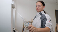 Ein Mädchen mit Brille und Arbeitskleidung streicht eine Wand mit einer Malerrolle weiß an.