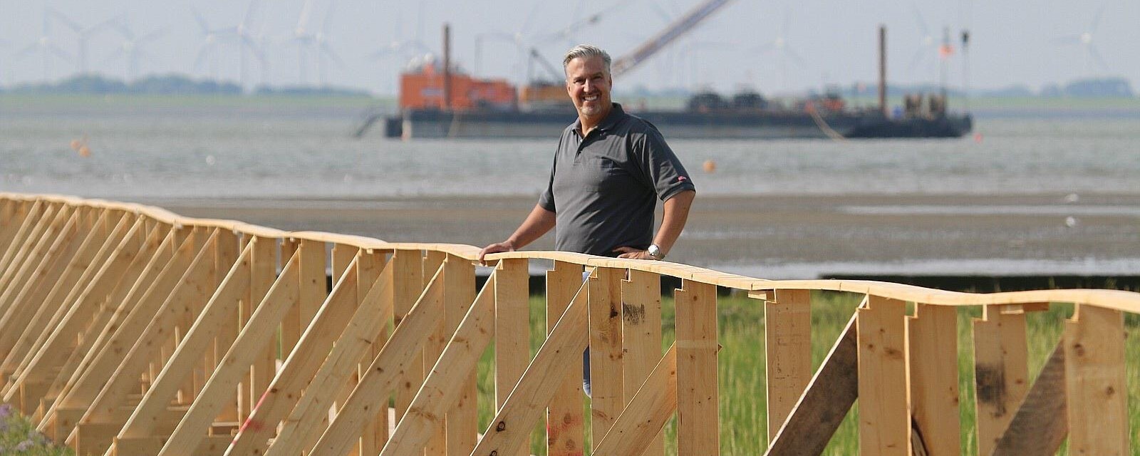 KI generiert: Ein Mann steht neben einer Holzstruktur vor einer Küstenlandschaft mit einem Schiff im Hintergrund.
