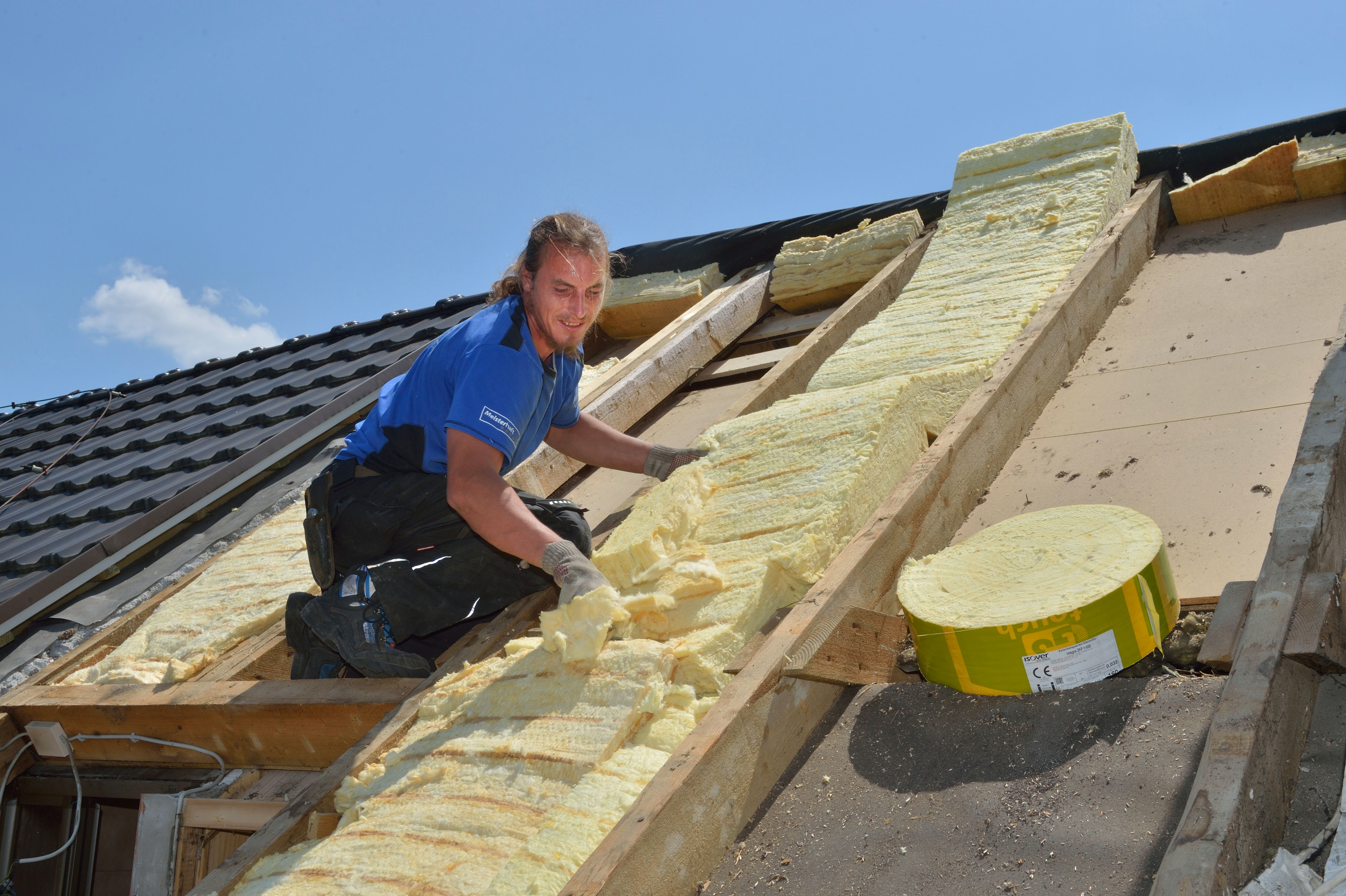 Ein Handwerker arbeitet auf einem Dach und hantiert mit gelbem Dämmmaterial – vermutlich zur Isolierung.