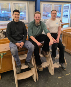 KI generiert: Drei Personen sitzen in einer Werkstatt auf hölzernen Treppenhockern.