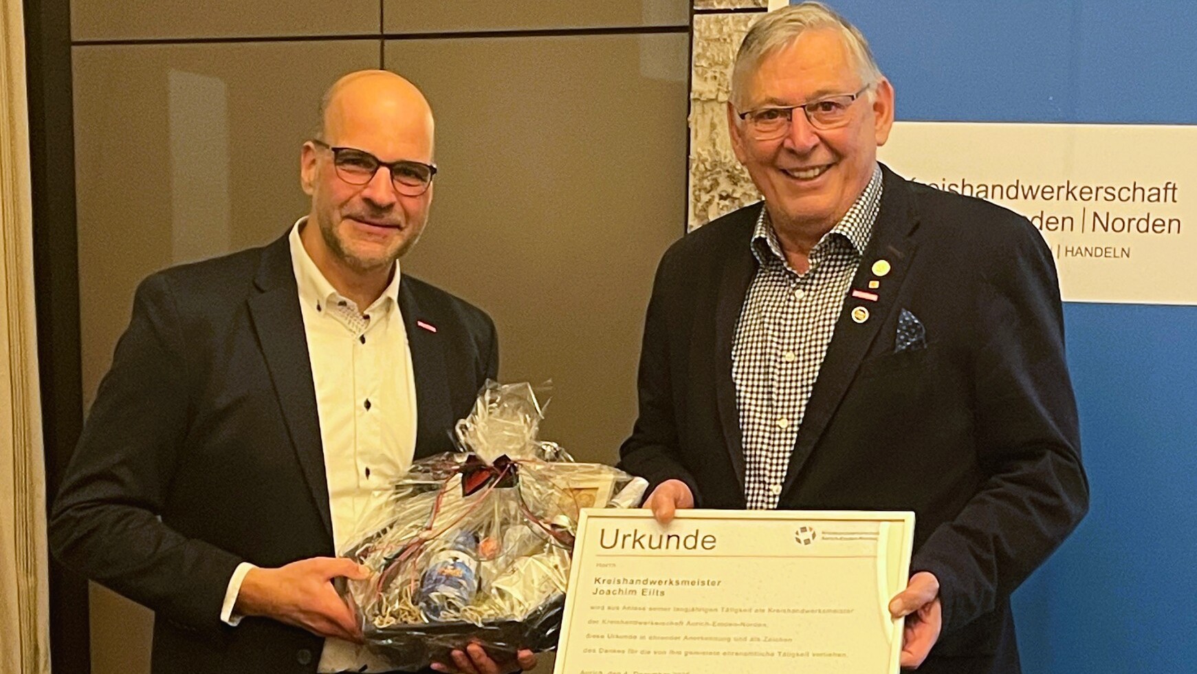 Der neue Kreishandwerksmeister Jens Boelen (l.) und sein Vorgänger Joachim Eilts posieren auf einem Bild mit einer Urkunde und einem Präsentkorb..