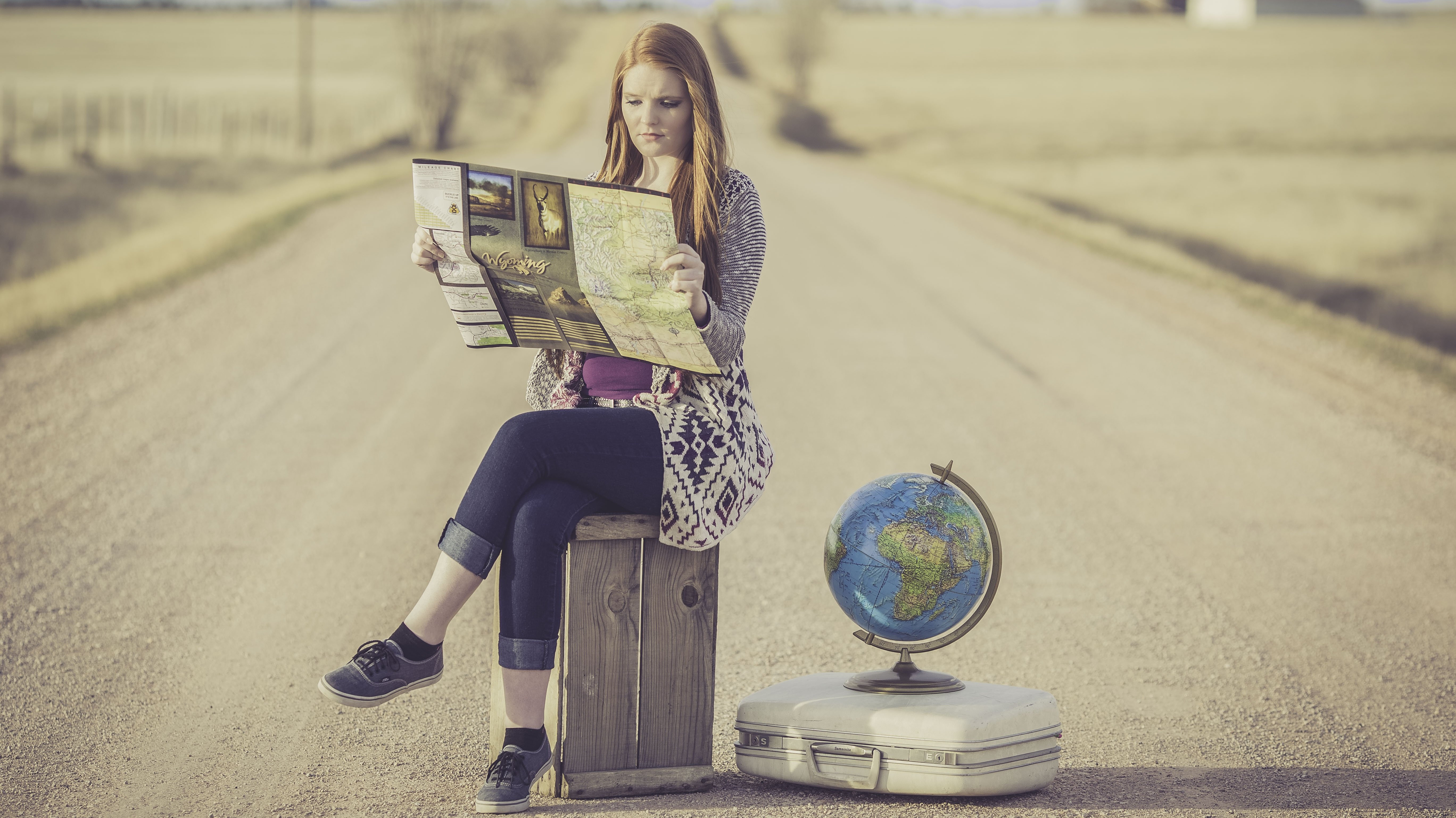 Eine Frau sitzt auf einer Kiste mitten auf der Straße, sie guckt auf eine Wegekarte. Neben ihr liegt ein Koffer mit einem Globus drauf.