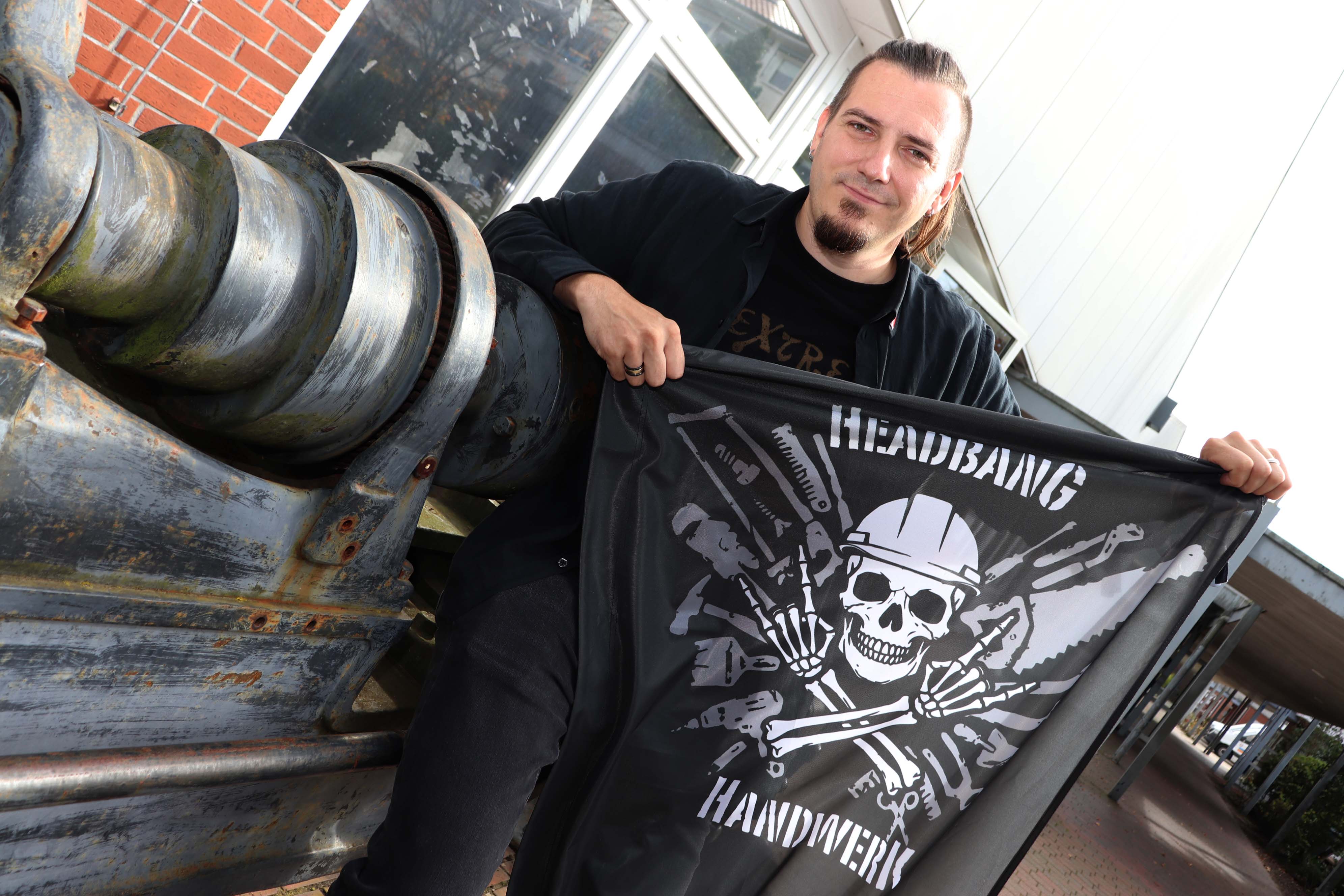 Der Hauptinhalt des Bildes ist ein Mann, der eine schwarze Flagge mit dem Aufdruck "HEADBANG HANDWERK" trägt.