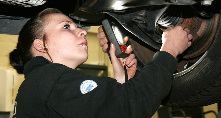 Eine Kfz-Mechatronikerin leuchtet mit einer Lampe unter einem aufgebockten Auto.
