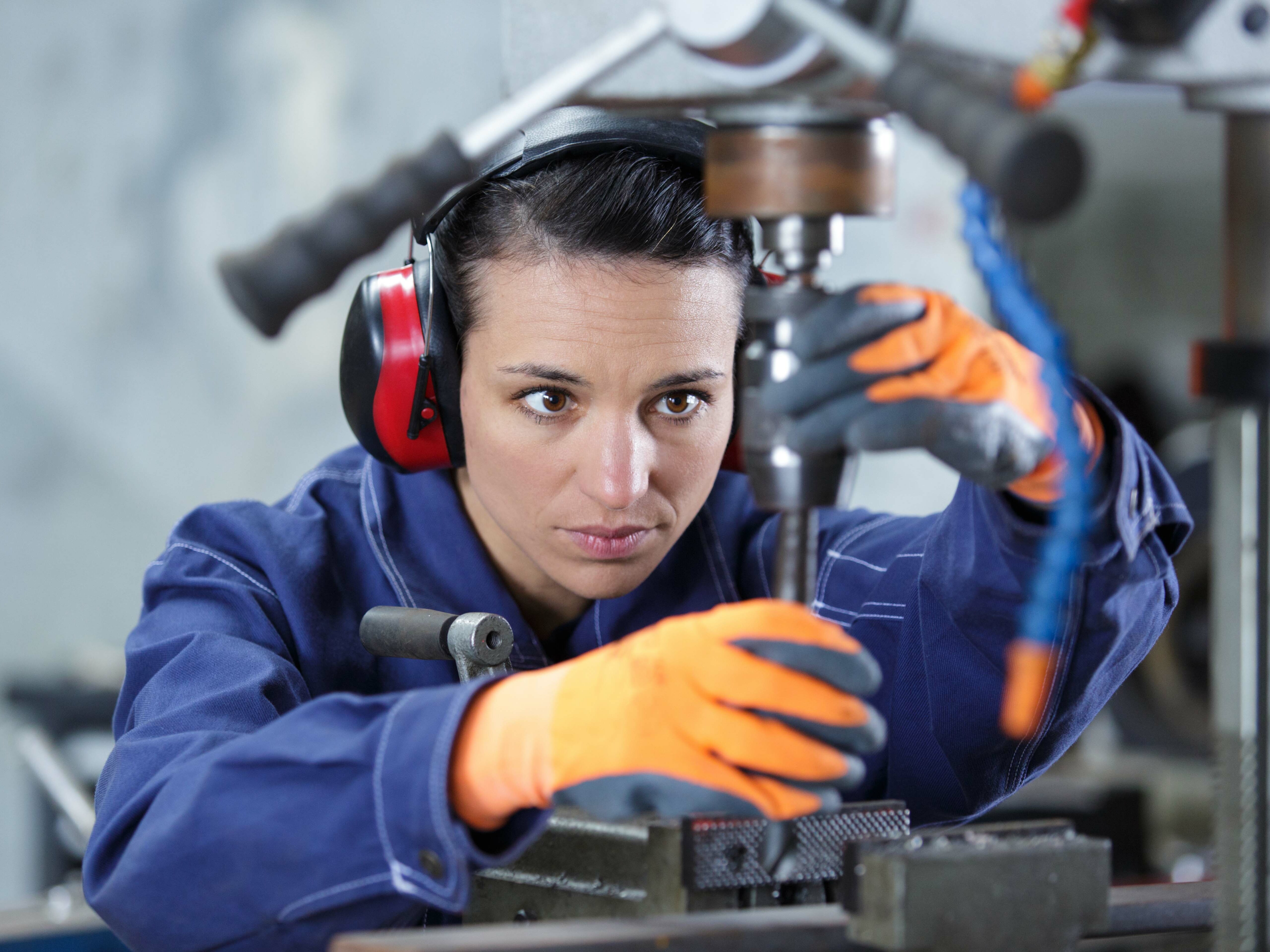 Eine Frau in Arbeitskleidung und Gehörschutz arbeitet konzentriert an einer Standbohrmaschine und hält ein Werkstück mit beiden Händen fest.