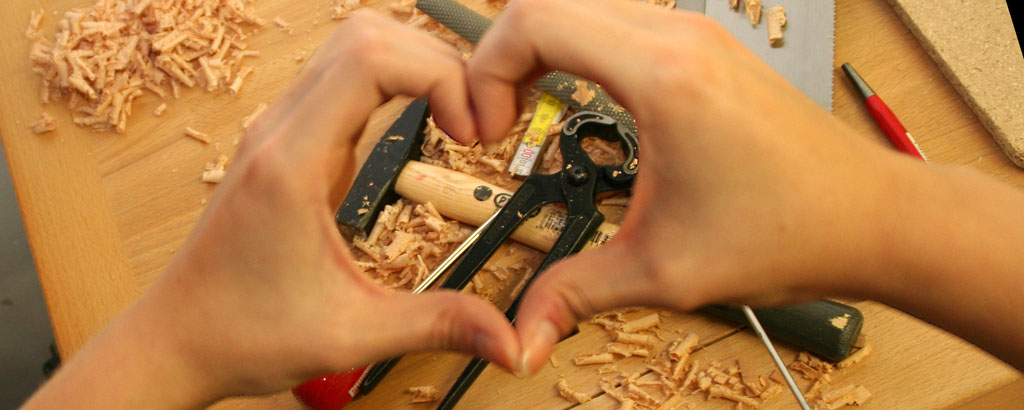 Mit der Hand wird ein Herz geformt, im Hintergrund liegen Werkzeuge.