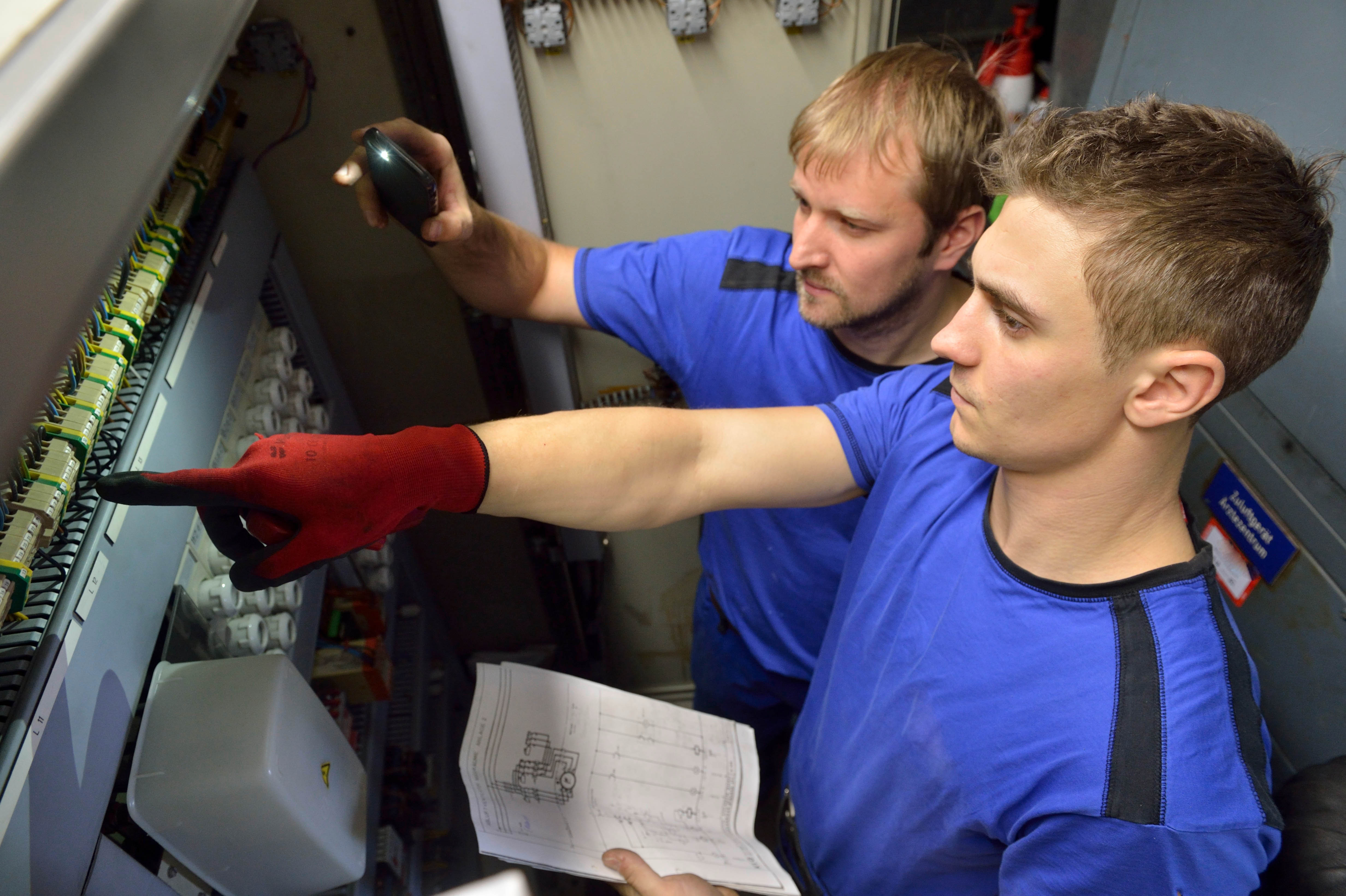 Zwei Männer in blauen Arbeitsshirts arbeiten an einem Schaltschrank. Einer hält einen Schaltplan, der andere zeigt mit einem roten Handschuh auf Bauteile – vermutlich eine technische Einweisung oder Wartung.