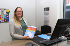 KI generiert: Eine Person steht an einem Schreibtisch und hält eine bunte Broschüre in der Hand.