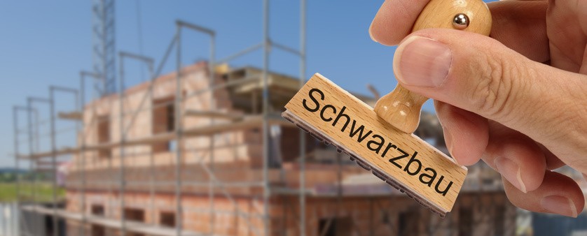 Ein Stempel mit der Aufschrift ‚Schwarzbau‘ im Vordergrund, dahinter eine Baustelle mit einem Haus im Rohbau.