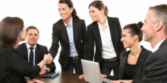 Sechs Geschäftspersonen in formeller Kleidung sitzen und stehen um einen Konferenztisch. Zwei Frauen in der Mitte geben sich die Hand, während die anderen lächelnd zusehen. Auf dem Tisch stehen ein Laptop und ein Notizbuch.