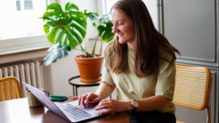 Eine frau mit braunen langen Haaren sitzt an einem Tisch und arbeitet an einem Laptop.