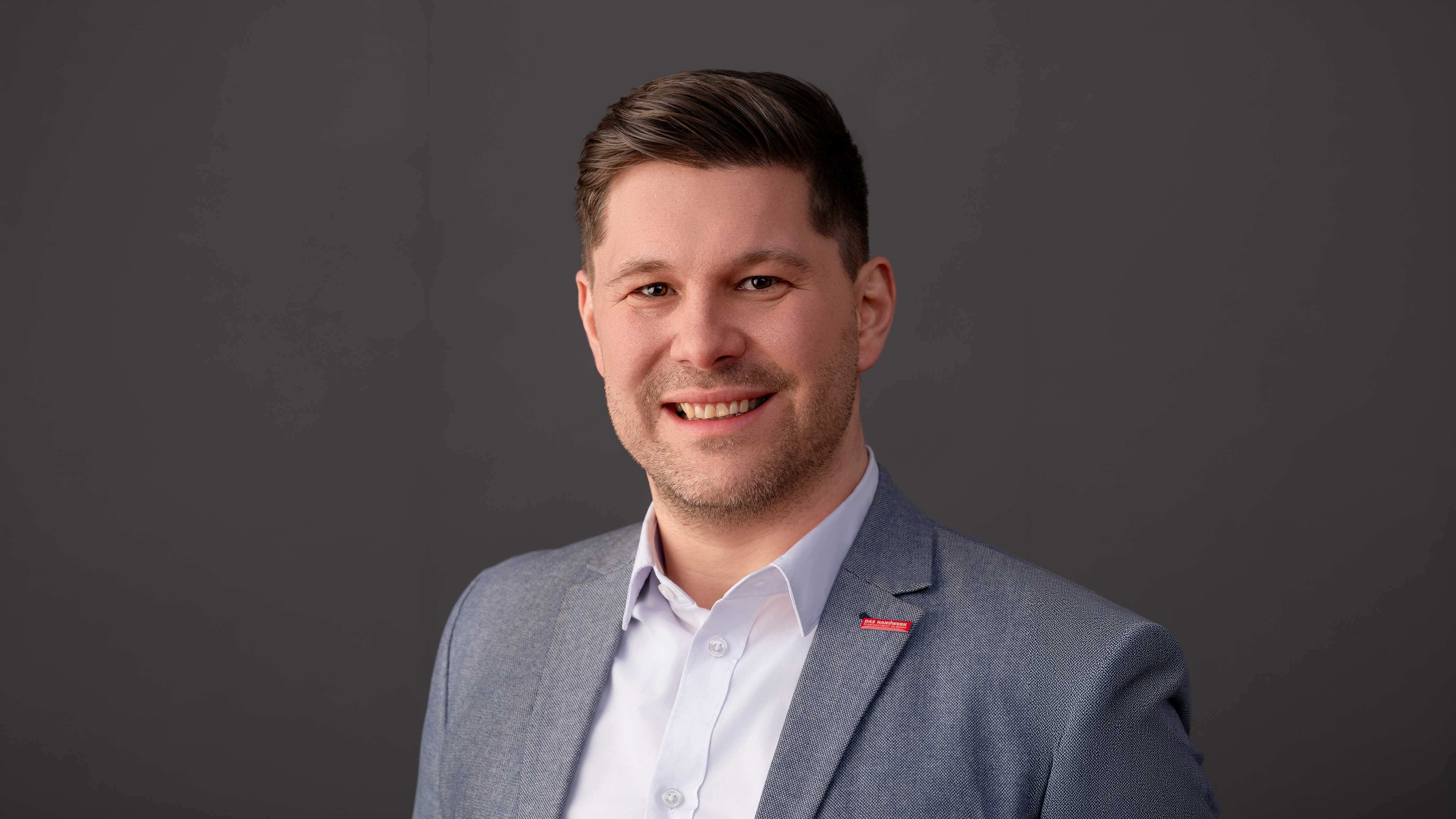 KI generiert: Es zeigt einen lächelnden Mann in einem grauen Anzug vor einem neutralen Hintergrund.