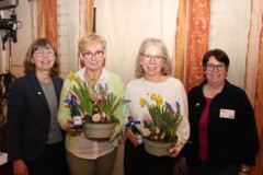 KI generiert: Das Bild zeigt vier Personen, von denen zwei Blumenarrangements und kleine Flaschen in den Händen halten, während sie lächeln.