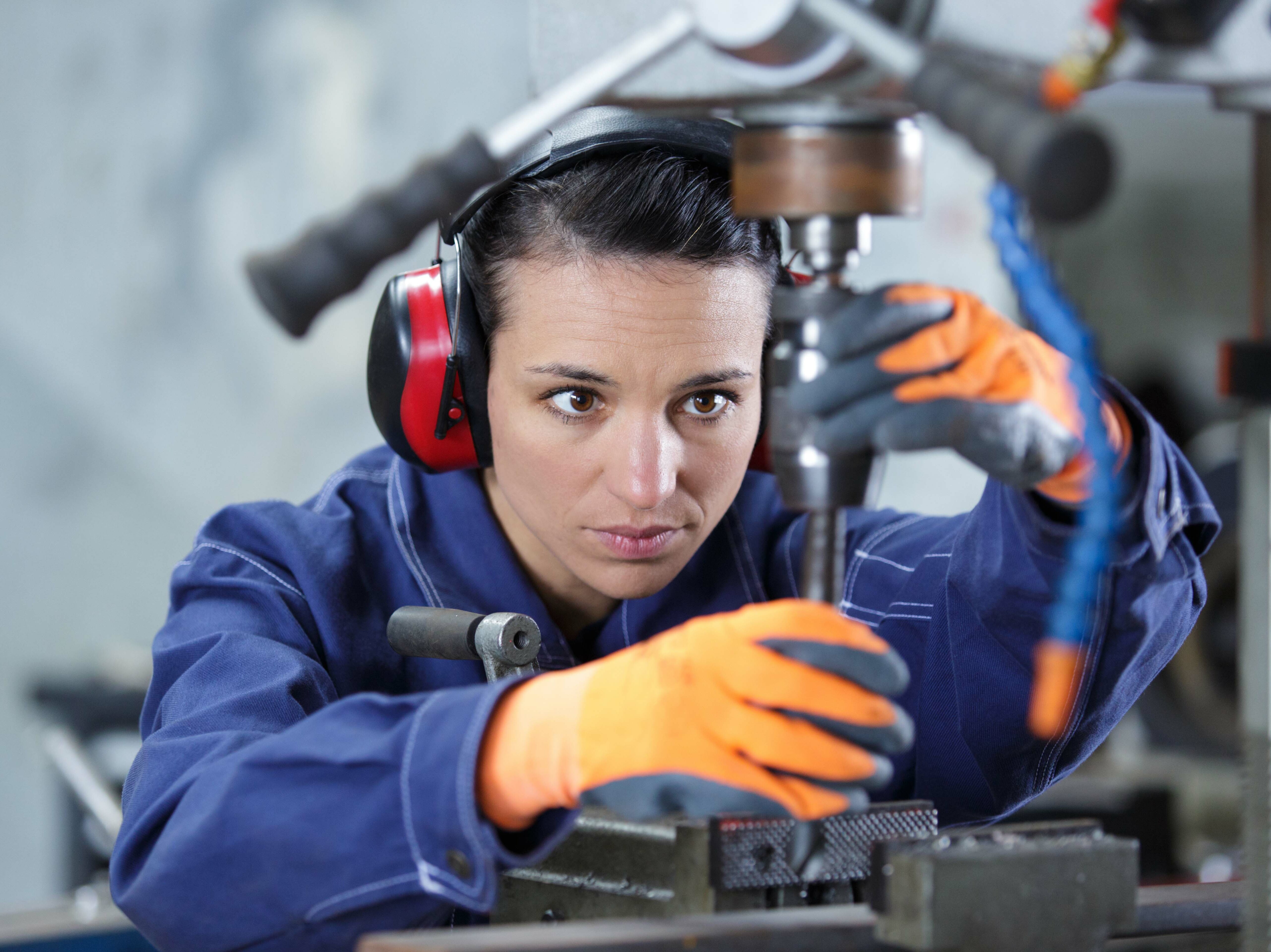 Eine Frau in Arbeitskleidung und Gehörschutz arbeitet konzentriert an einer Standbohrmaschine und hält ein Werkstück mit beiden Händen fest.
