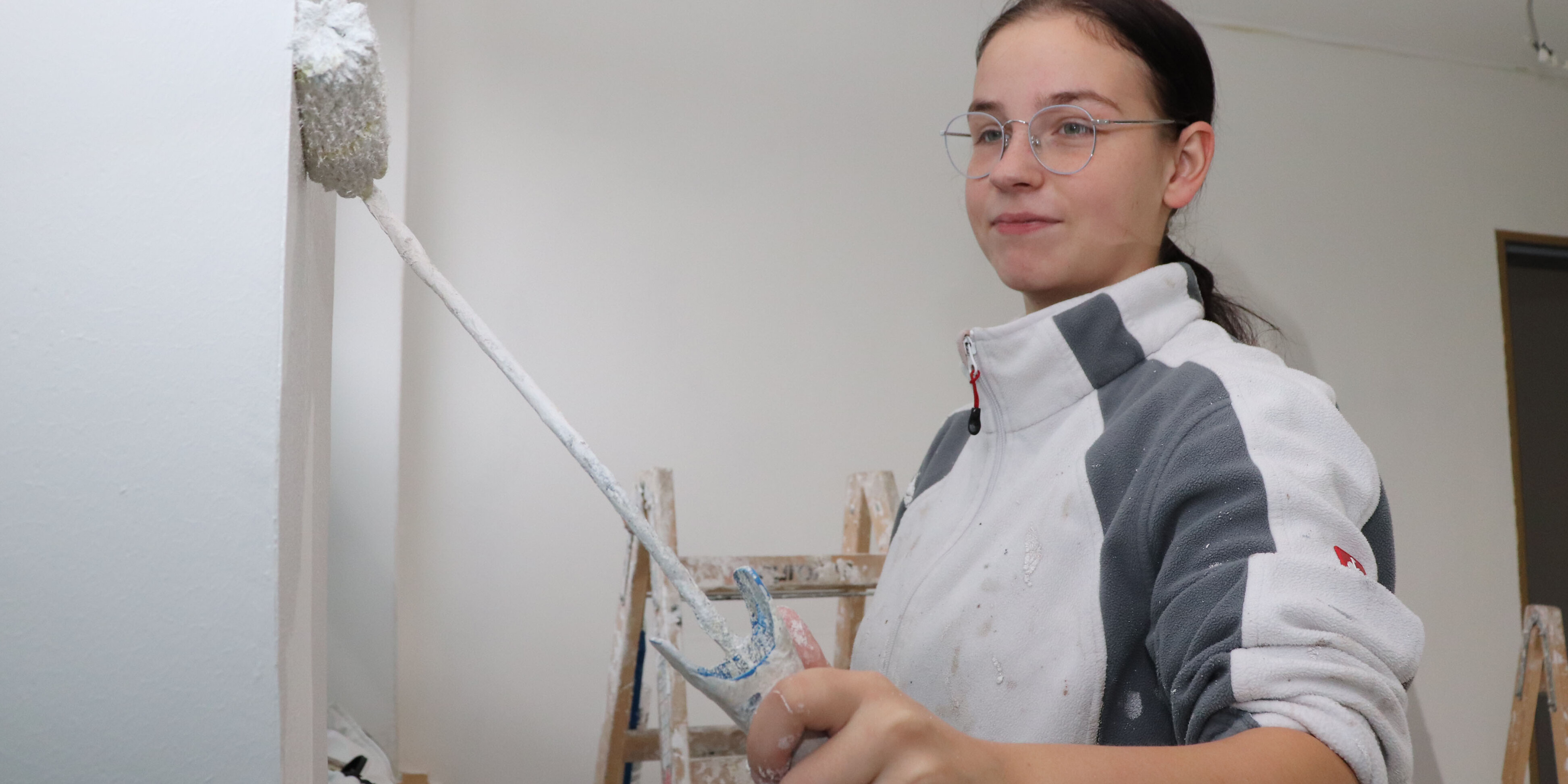Ein Mädchen mit Brille und Arbeitskleidung streicht eine Wand mit einer Malerrolle weiß an.